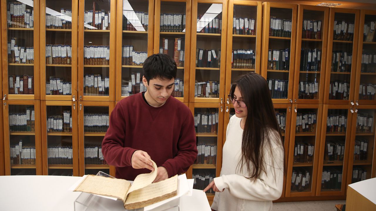 Imagen de estudiantes mirando un libro. Al fonde se ve una vitrina con libros